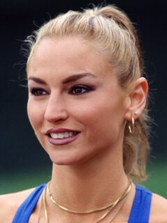 Адриана Ла Серва с собранными в хвост светлыми волосами, в синем топе и золотых украшениях.