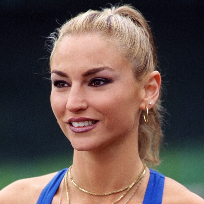 Адриана Ла Серва с собранными в хвост светлыми волосами, в синем топе и золотых украшениях.