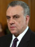 Джонни Сэк с зачёсанными назад седыми волосами, в тёмном костюме и фиолетовом галстуке.