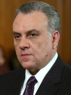 Джонни Сэк с зачёсанными назад седыми волосами, в тёмном костюме и фиолетовом галстуке.